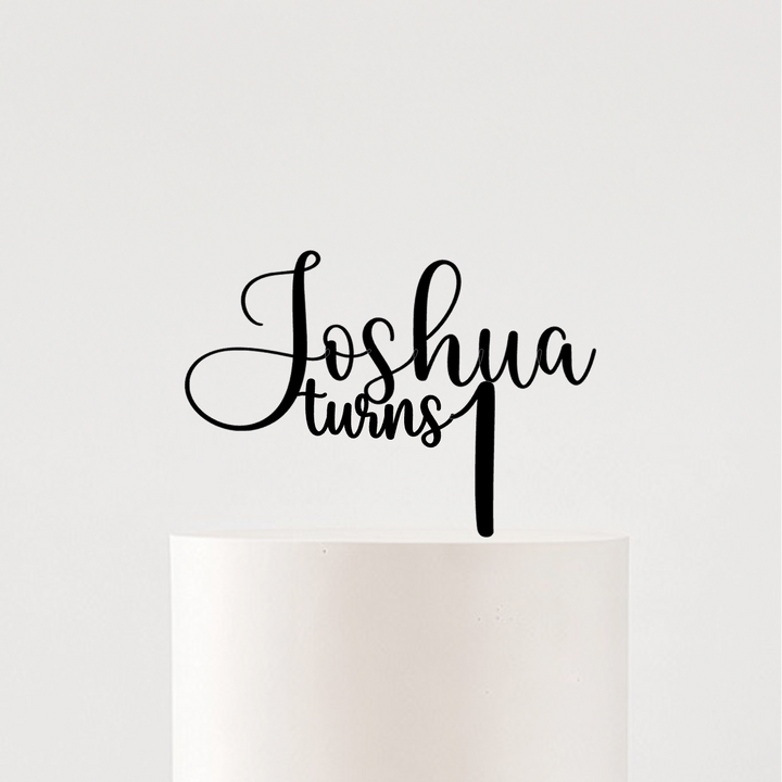 A white frosted cake on a white pedestal stand with a large sheer white bow around the base, topped with a black script cake topper that reads &
