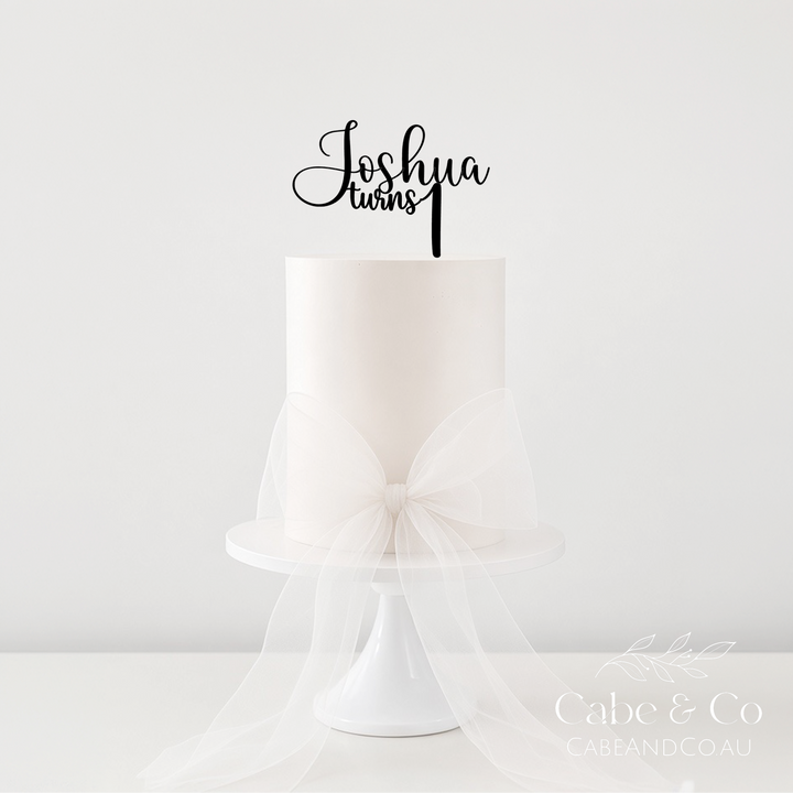 A white frosted cake on a white pedestal stand with a large sheer white bow around the base, topped with a black script cake topper that reads &