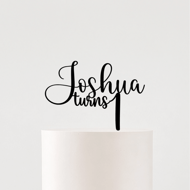 A white frosted cake on a white pedestal stand with a large sheer white bow around the base, topped with a black script cake topper that reads &