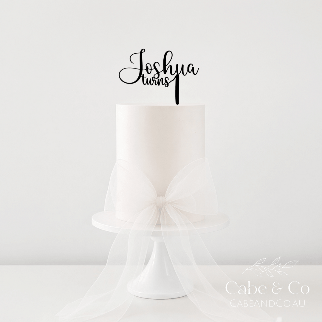A white frosted cake on a white pedestal stand with a large sheer white bow around the base, topped with a black script cake topper that reads &