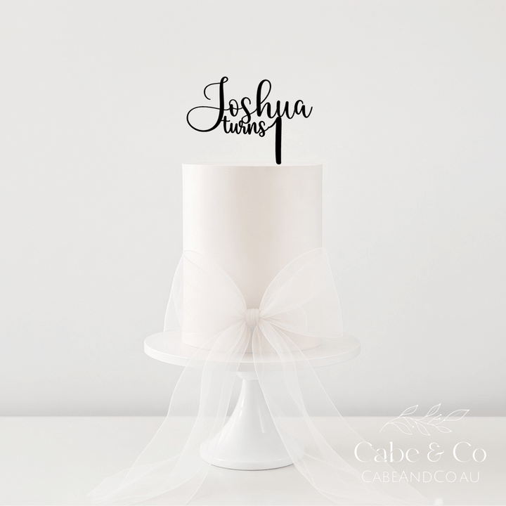 A white frosted cake on a white pedestal stand with a large sheer white bow around the base, topped with a black script cake topper that reads &