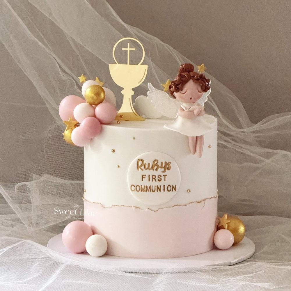 A decorated round cake for a first communion celebration, featuring a white top half with gold lettering reading 'Ruby's First Communion,' a gold chalice topper with a cross, a small angel figurine, pastel pink and white balloons, and gold star accents. The cake sits on a white board with tulle fabric in the background.