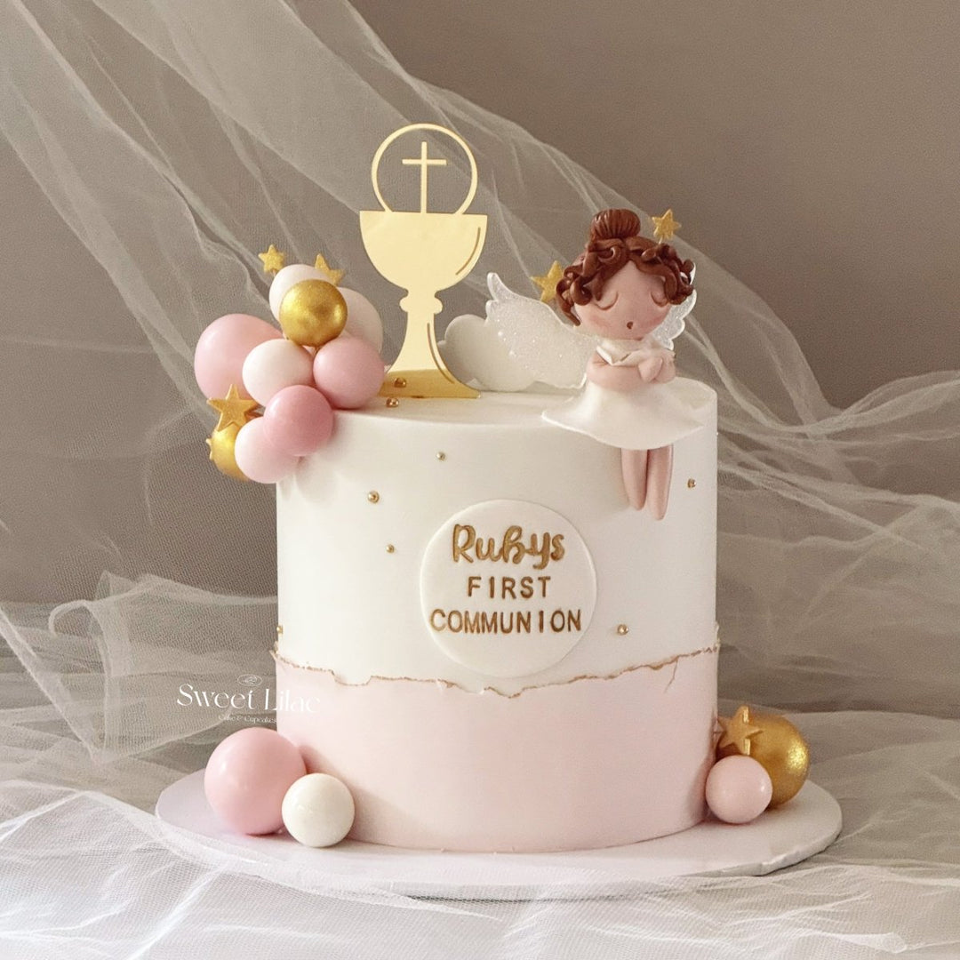 A decorated round cake for a first communion celebration, featuring a white top half with gold lettering reading &