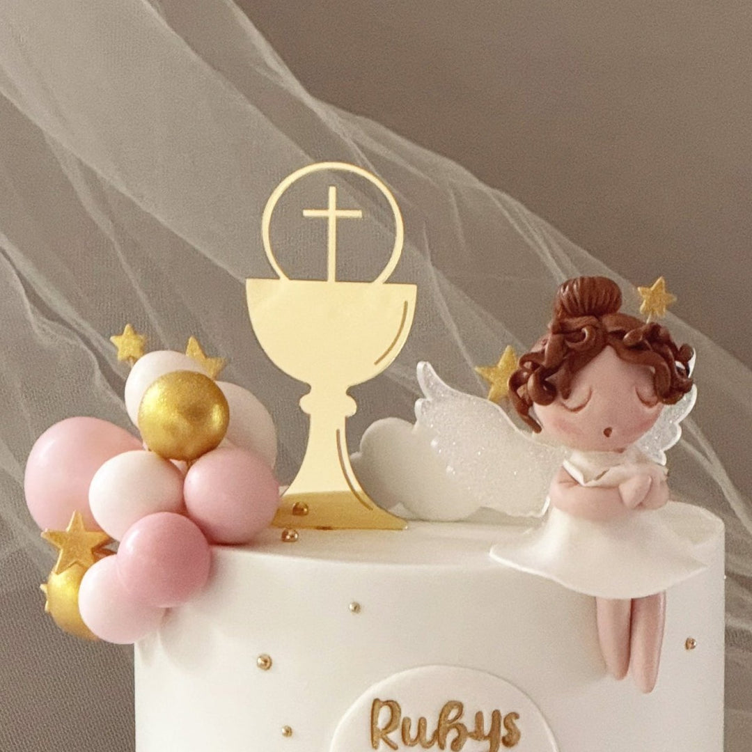 A decorated first communion cake with a white and pale pink finish, gold lettering reading &
