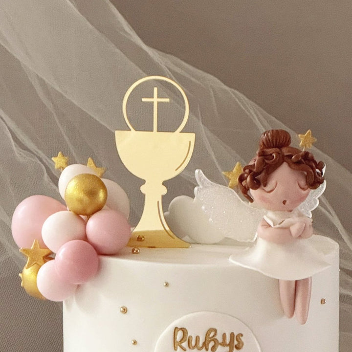 A decorated first communion cake with a white and pale pink finish, gold lettering reading &