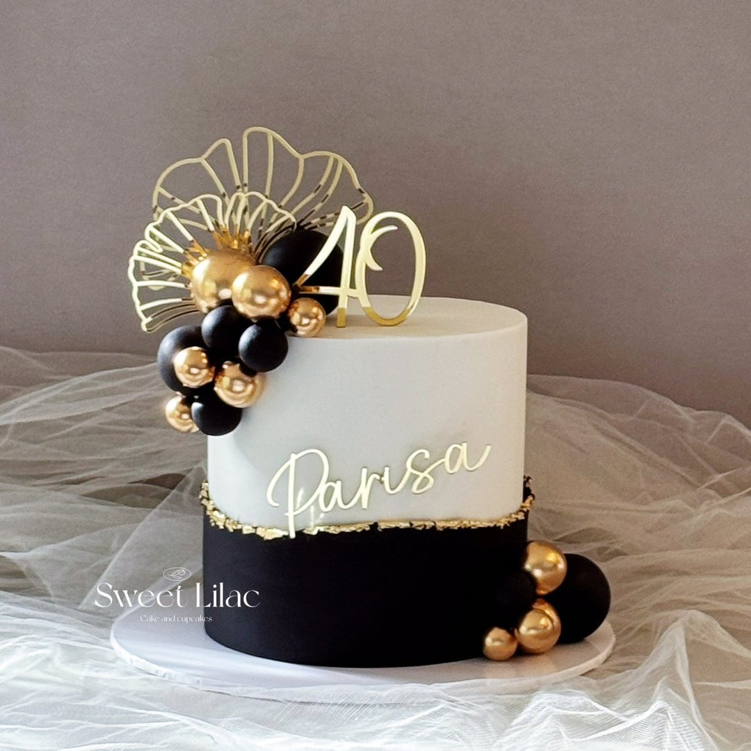 A custom 40th birthday cake with a white top half, a black bottom half with gold glitter trim, and gold and black balloon decorations. The cake is decorated with a gold &