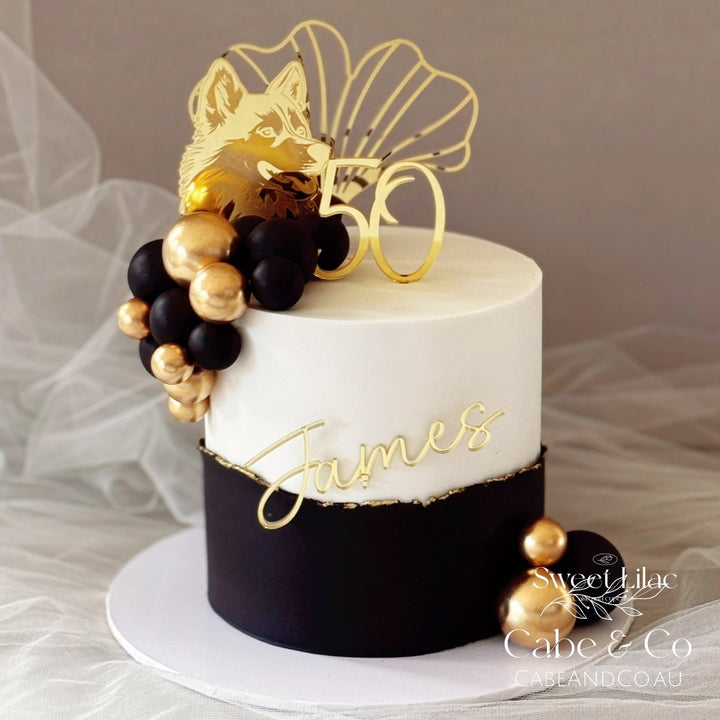 A custom two-tier birthday cake with white and black frosting, decorated with gold foil script reading &