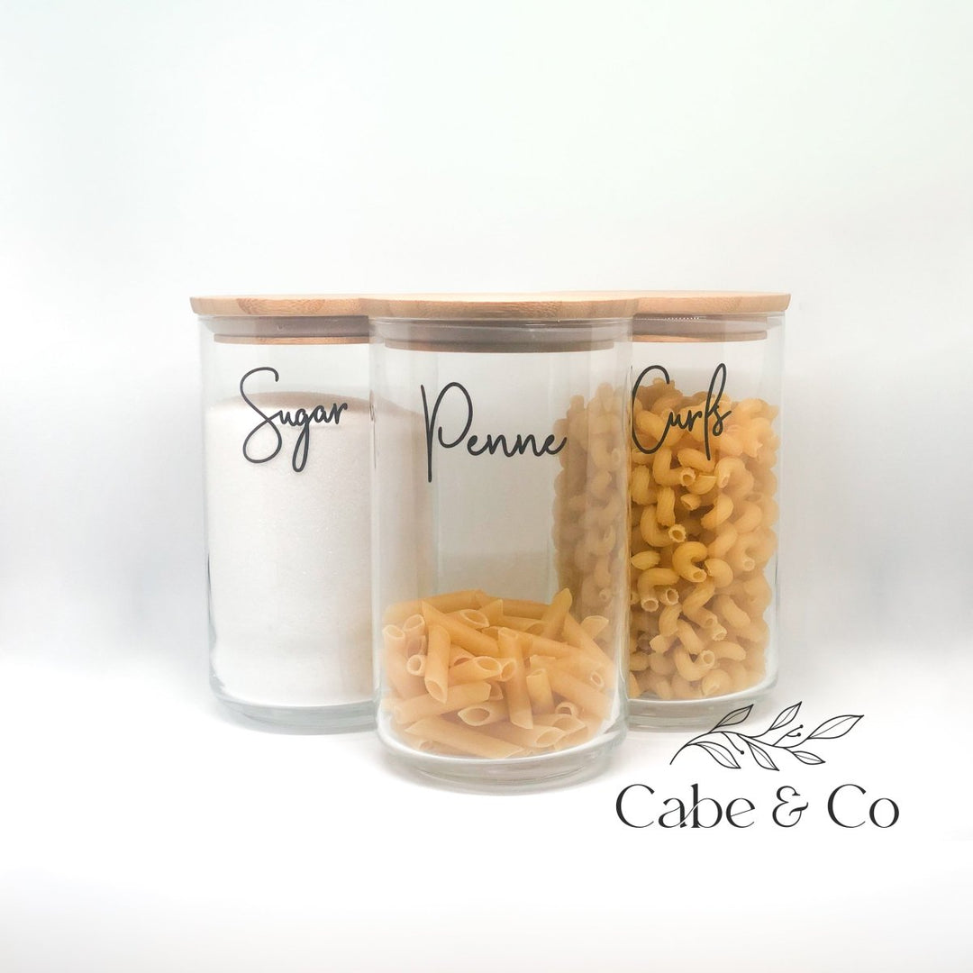 Three clear glass canisters with light wood lids, labeled &