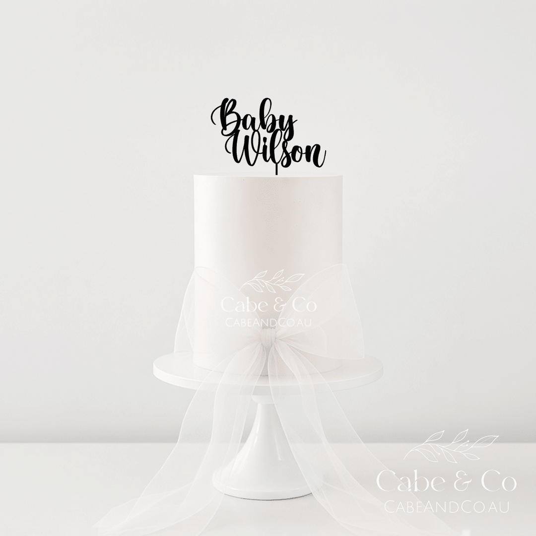 A single-tier white cake on a white pedestal stand with a sheer white tulle bow around the waist, topped with a black script cake topper reading &