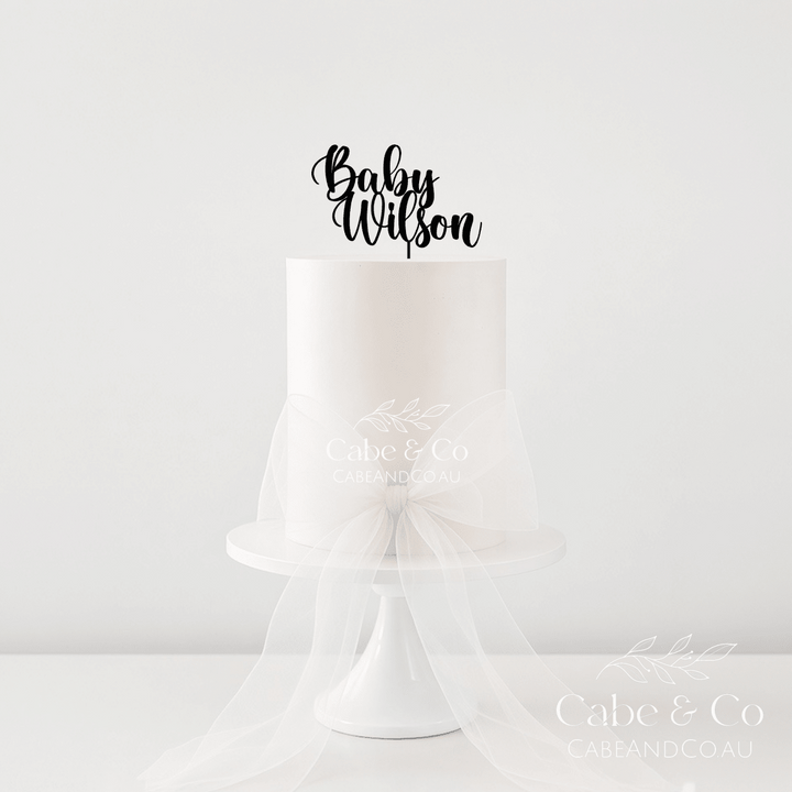 A single-tier white cake on a white pedestal stand with a sheer white tulle bow around the waist, topped with a black script cake topper reading &