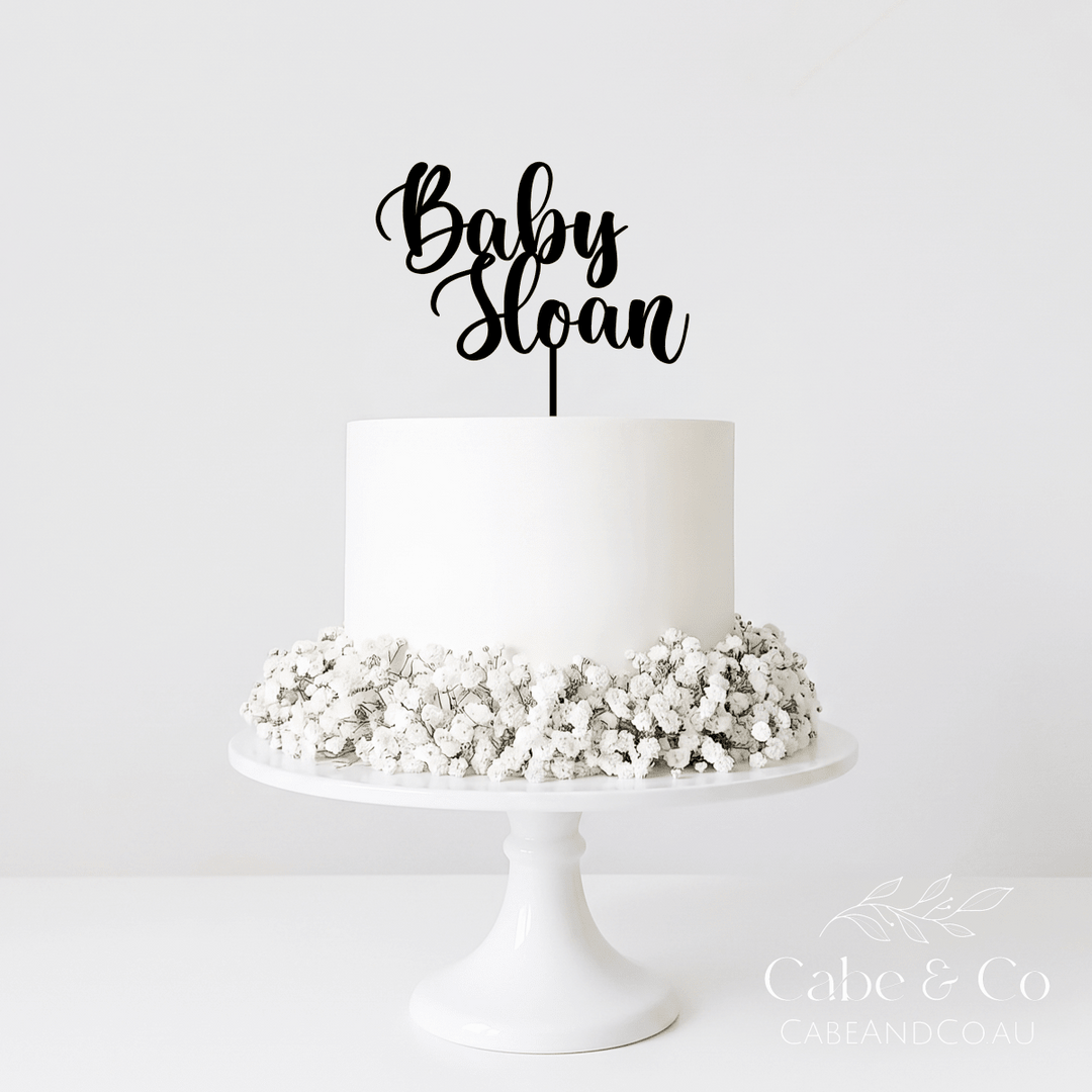 A white frosted cake on a white pedestal stand, decorated with white baby&