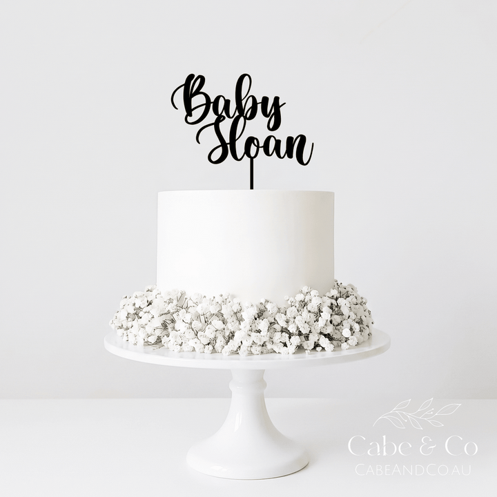 A white frosted cake on a white pedestal stand, decorated with white baby&