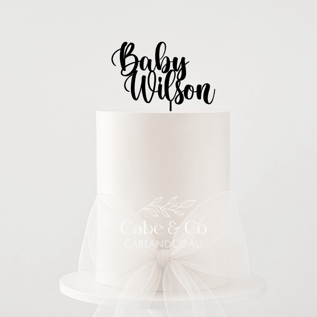 A single-tier white cake on a white pedestal stand with a sheer white tulle bow around the waist, topped with a black script cake topper reading &