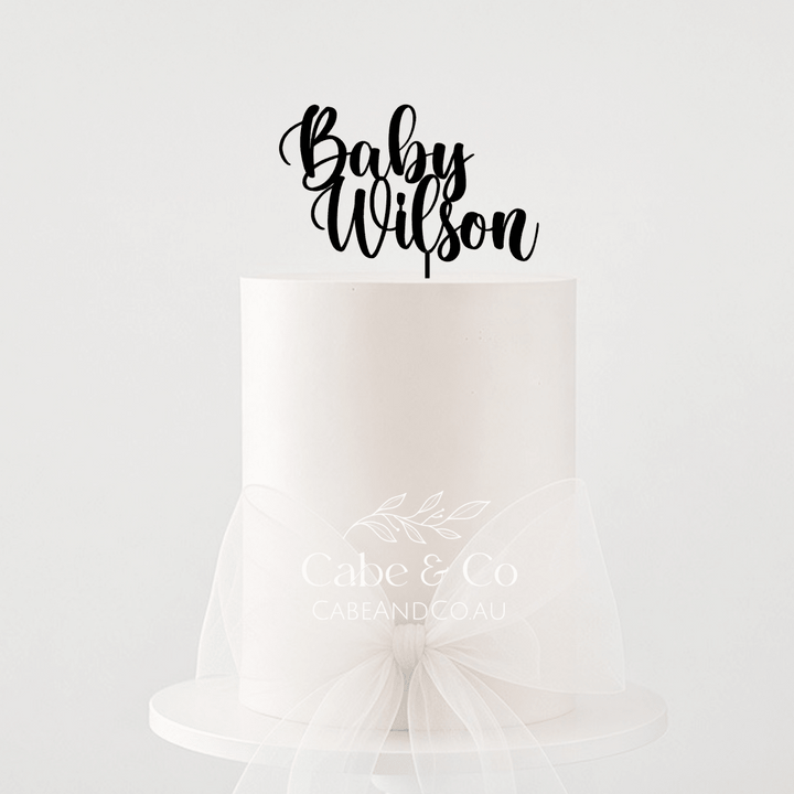 A single-tier white cake on a white pedestal stand with a sheer white tulle bow around the waist, topped with a black script cake topper reading &