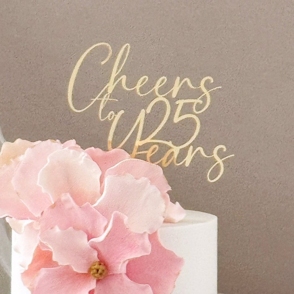 A close-up of a white cake decorated with large pink flower petals and a gold script cake topper that reads 'Cheers to 25 Years,' set against a taupe background with a sheer white fabric drape.