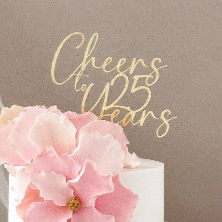 A close-up of a white cake decorated with large pink flower petals and a gold script cake topper that reads &