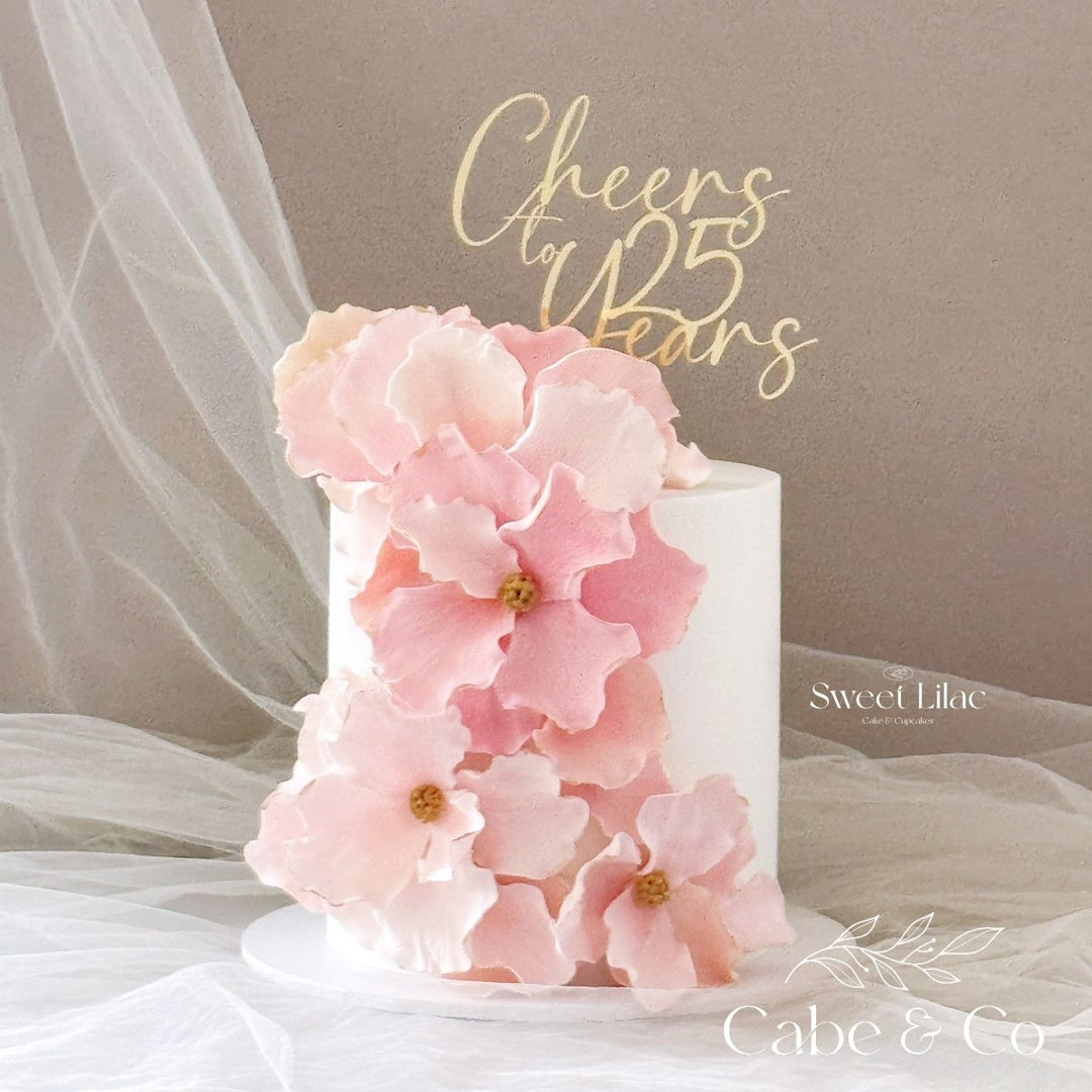 A white cylindrical cake decorated with large pale pink sugar flowers and a gold &
